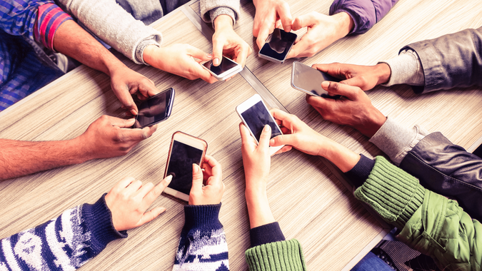 Hands holding phones in a circle of people. Fingers visible as they use their devices, with a focus on the phones rather than the individuals.